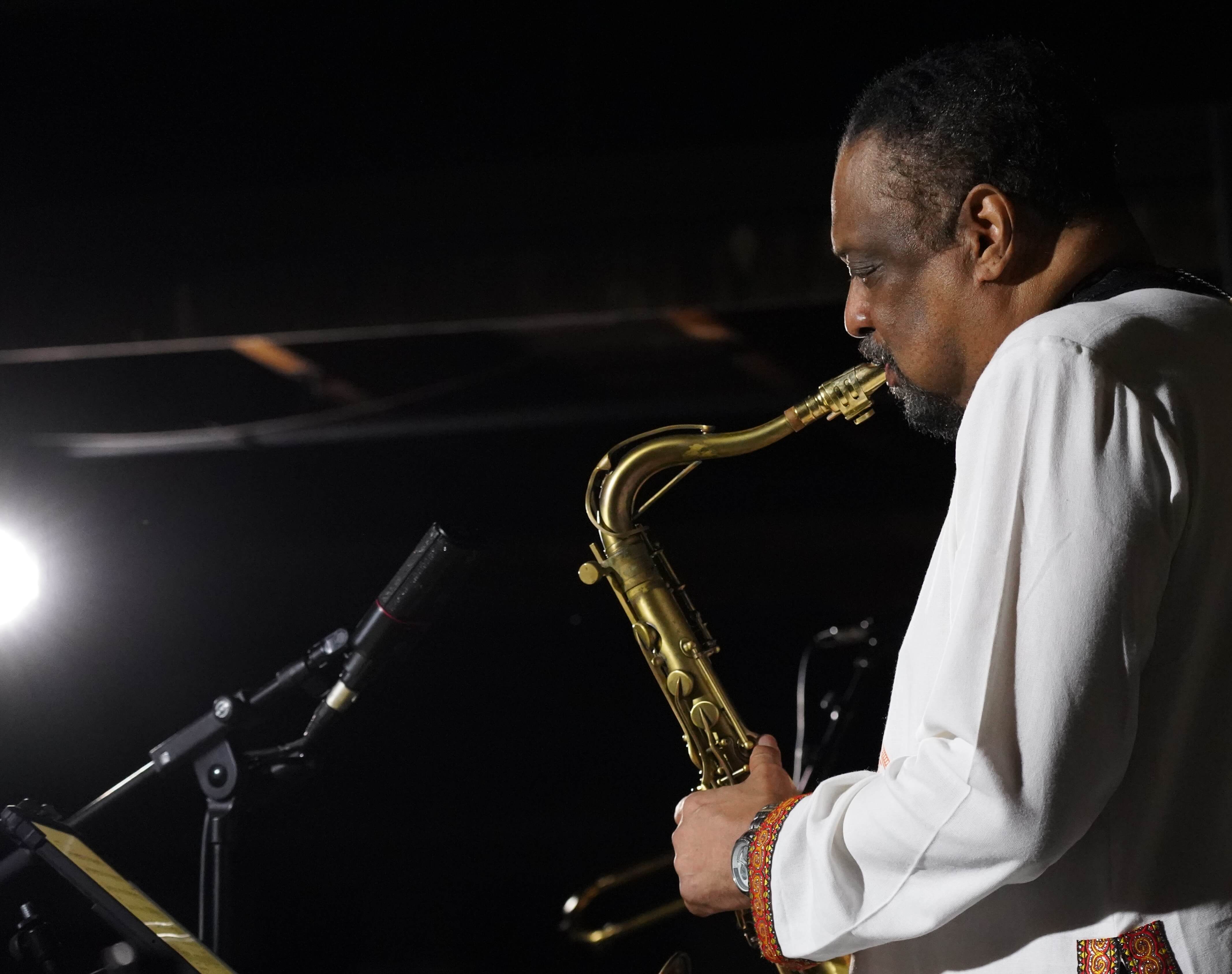 Tenorsaxophonist im weißen Hemd am Mikrofon, von hinten beleuchtet auf dunkler Bühne.