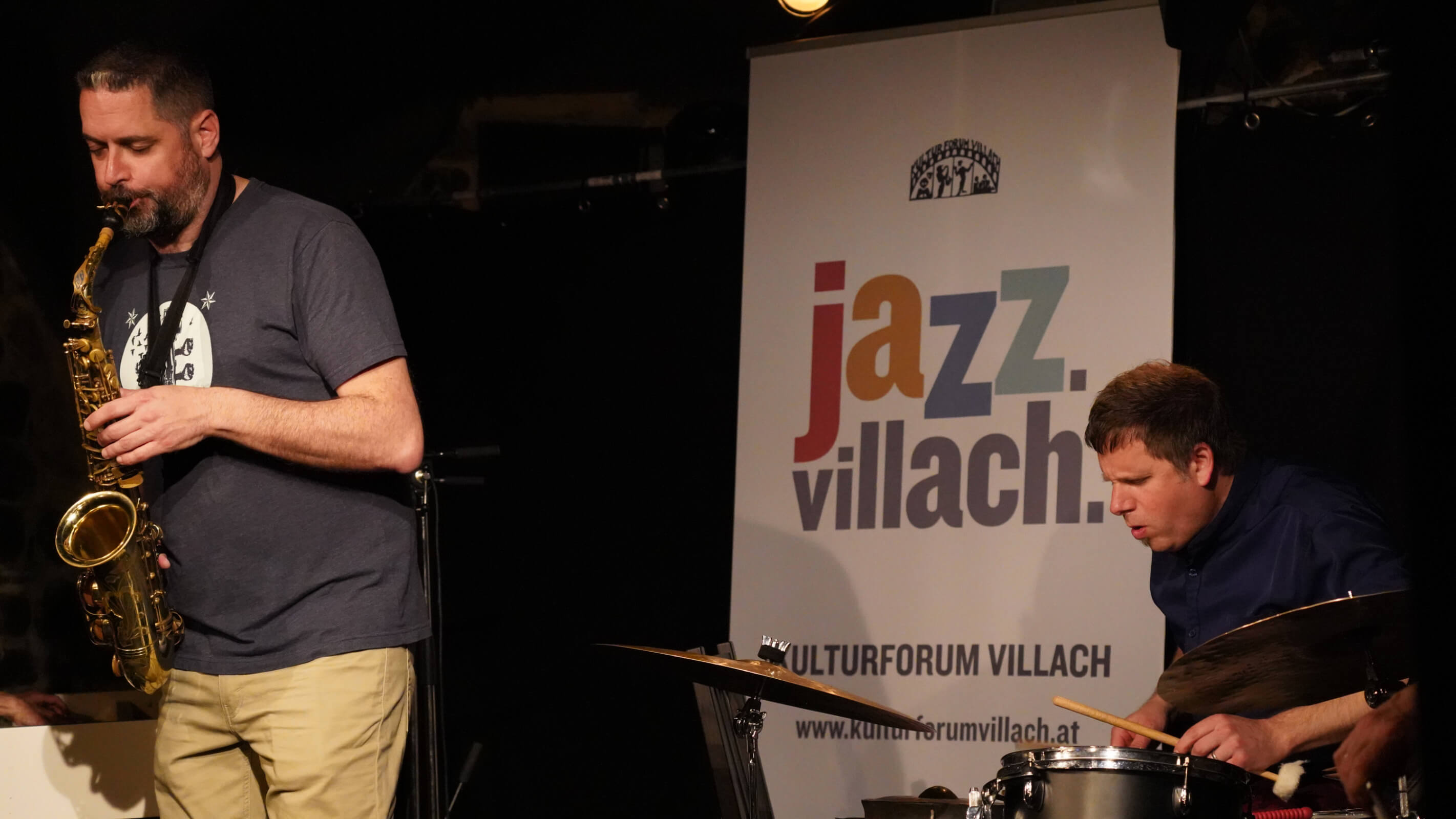 Altsaxophonist und Schlagzeuger gemeinsam auf der Bühne neben Jazz-Villach-Banner.