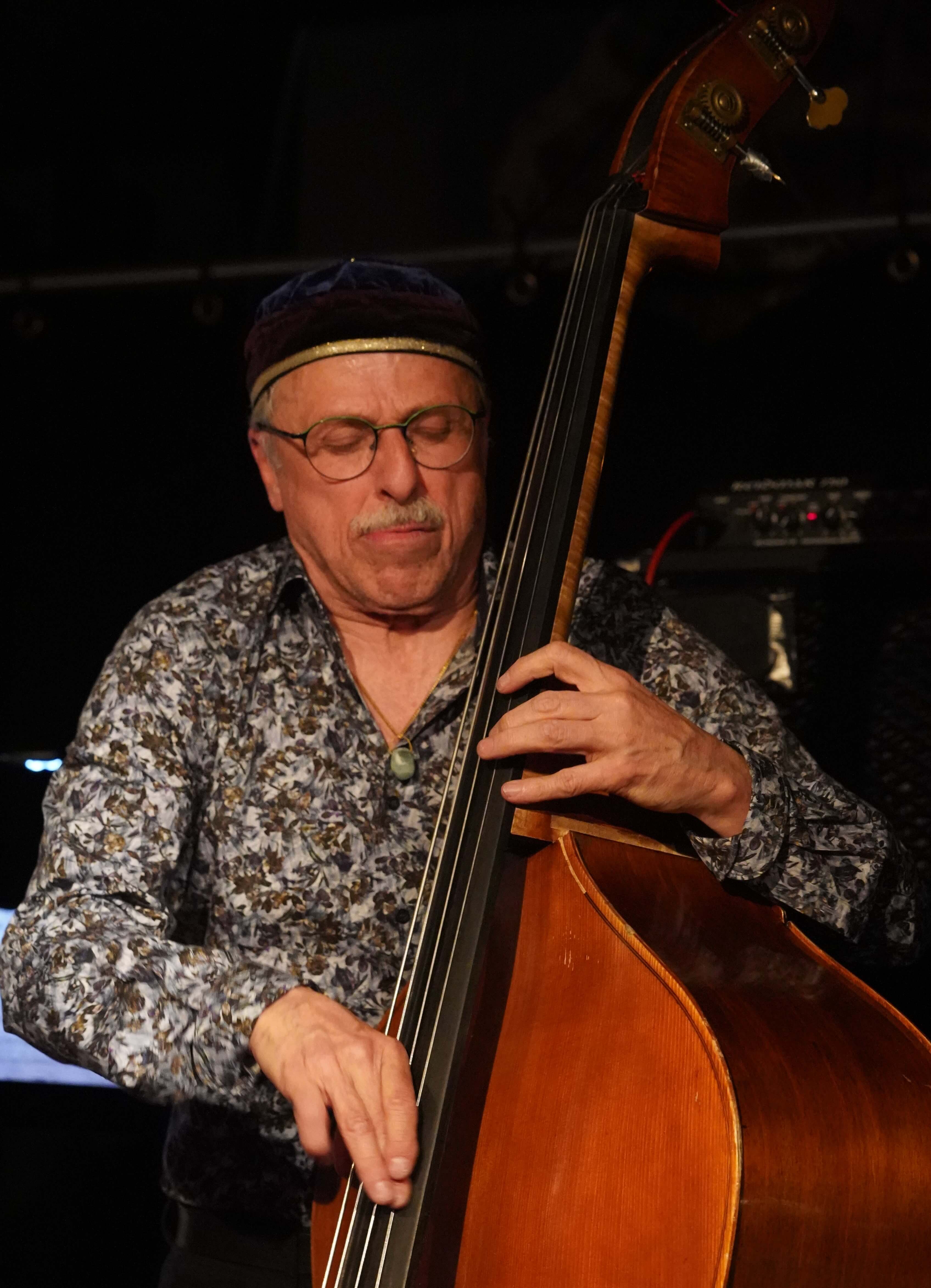 Kontrabassist im gemusterten Hemd auf dunkel beleuchteter Bühne.