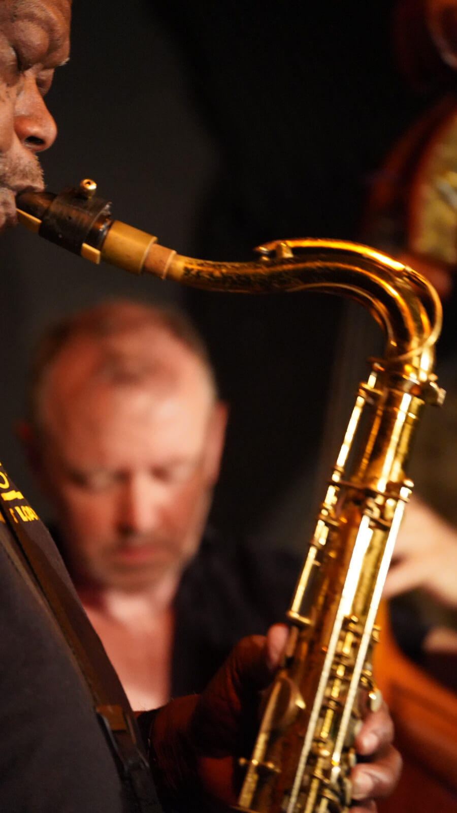 Nahaufnahme eines Tenorsaxophonisten, dahinter unscharfer Bassist auf dunkler Bühne.