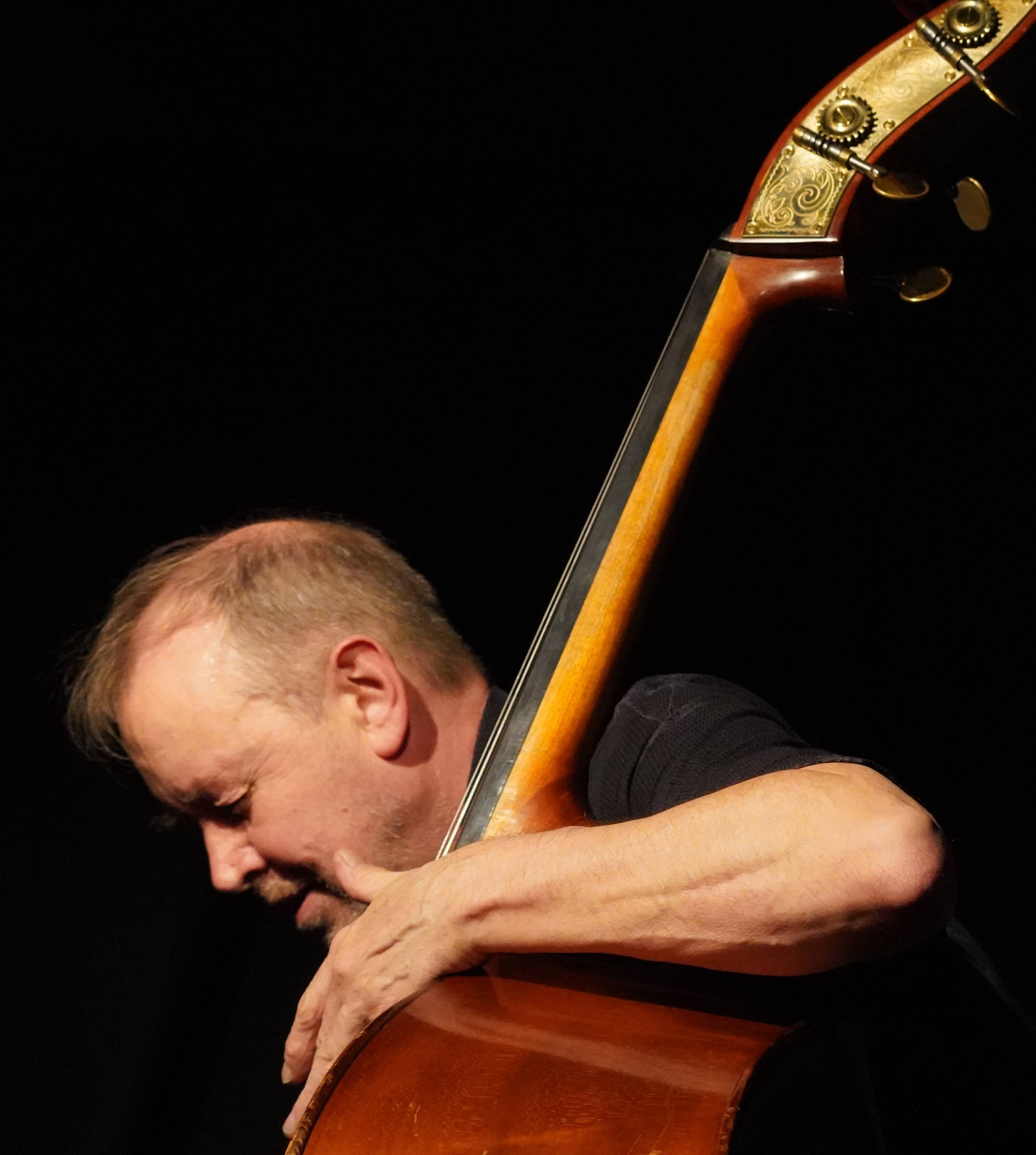 Kontrabassist auf der Bühne, Gesicht teils hinter dem Bass vor dunklem Hintergrund.