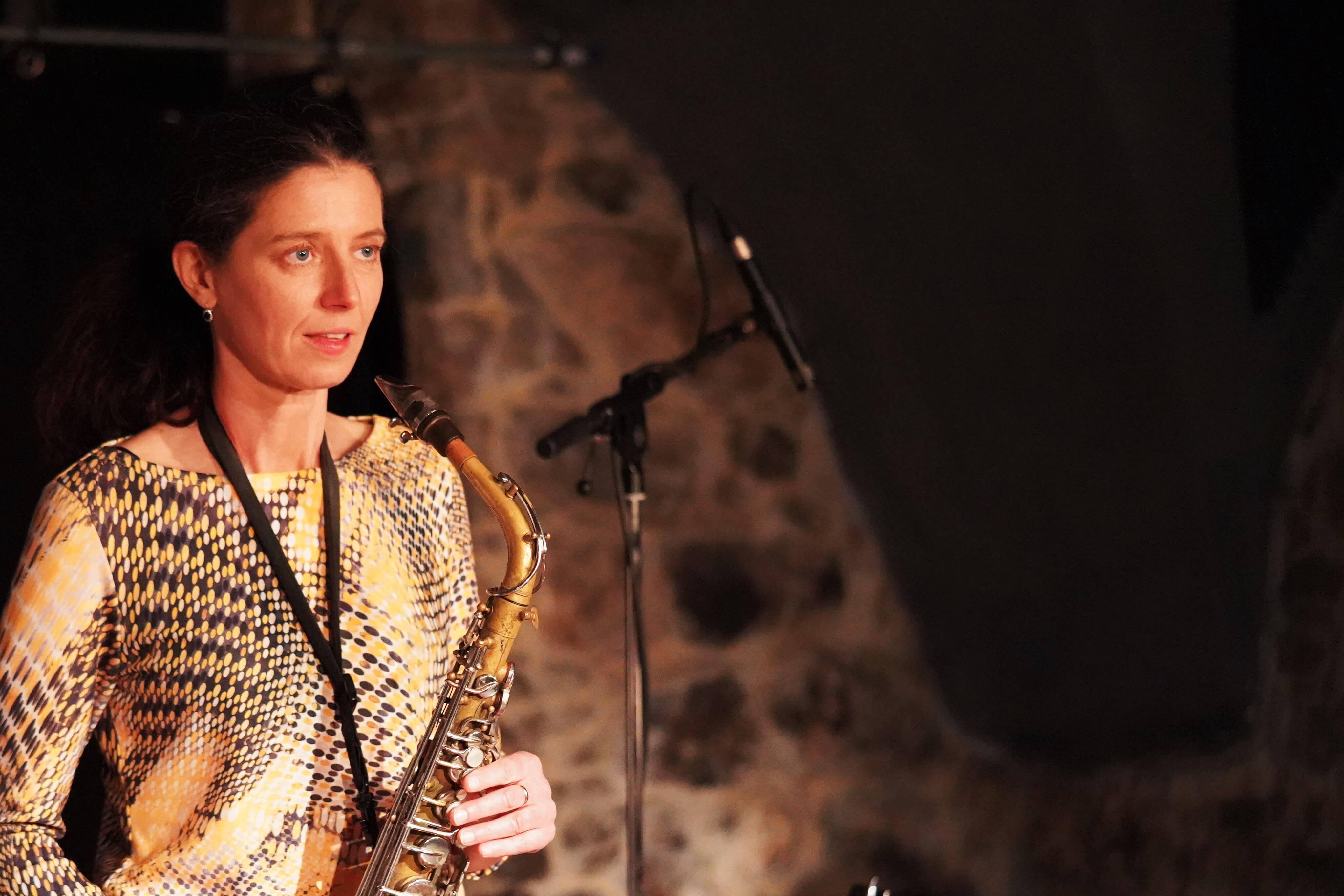 Altsaxophonistin im gemusterten Kleid vor Steinwand und Mikrofonen.