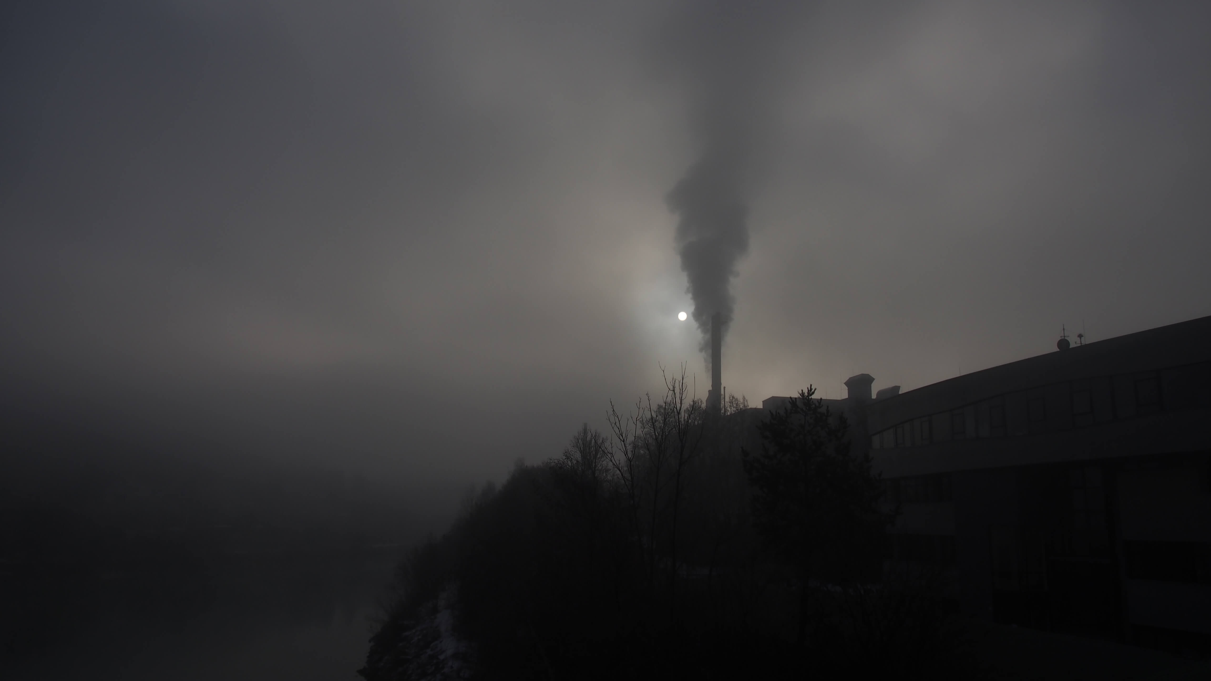 Industrieschornstein stößt Rauch in den diesigen Himmel, Sonne schwach durch den Nebel sichtbar.