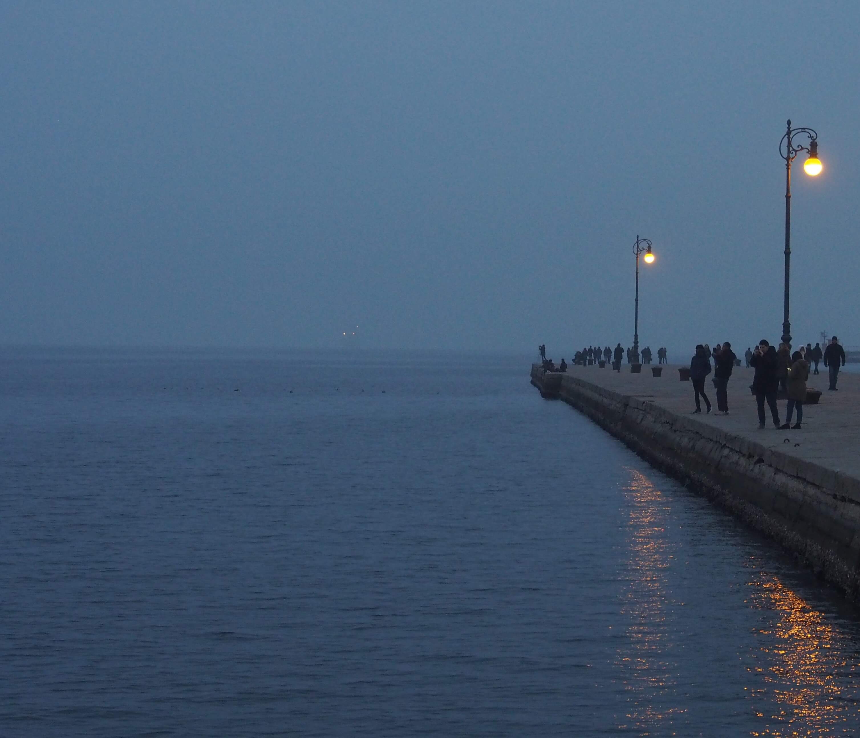 Meerespromenade im dichten Abendnebel, leuchtende Straßenlaternen und Menschen als Silhouetten am Wasser.